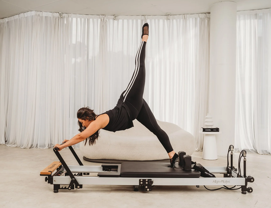 Woman practicing Pilates on a reformer machine beside a mat in a bright studio