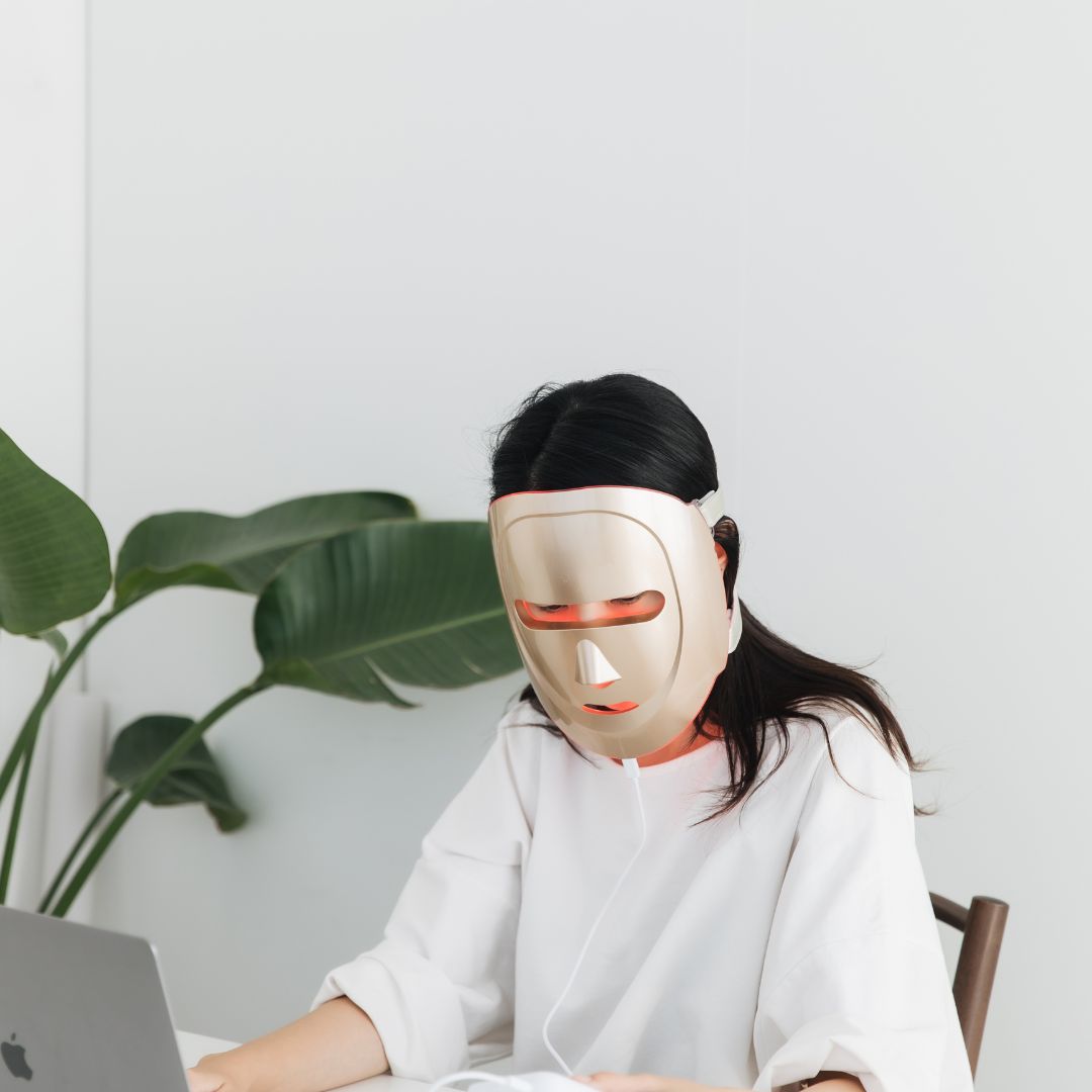 Person wearing a LED face mask in a home office setting with a plant in the background